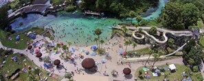An aerial view of a beach area with a roller coaster, clear turquoise water, and numerous umbrellas and people.