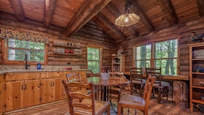 L’intérieur d’un chalet rustique, avec des murs en bois, une table à manger, des chaises et un coin cuisine doté d’étagères et d’une fenêtre.