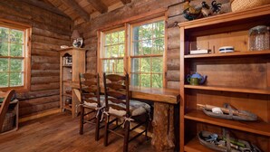 L’intérieur d’un chalet rustique en bois, avec une table à manger, des chaises, une bibliothèque et des fenêtres donnant sur un paysage verdoyant.