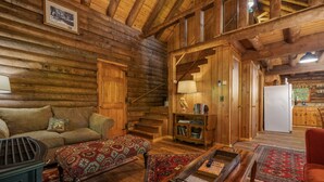 L’intérieur chaleureux d’un chalet en bois comprend un canapé, un pouf à motifs, une table basse en bois et un escalier menant à un coin cuisine.