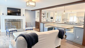 A modern living room with a fireplace, seating area, and a kitchen visible through an open doorway. - Spacious Home Near Wallis Sands Beach in Rye NH (Rye)