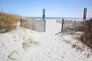 A sandy beach path leading to the ocean, flanked by wooden fences and sparse vegetation.