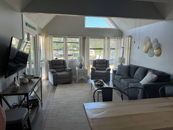 A spacious living room with a large window, two grey armchairs, a grey sofa, a TV mounted on the wall, and a small table with a lamp.