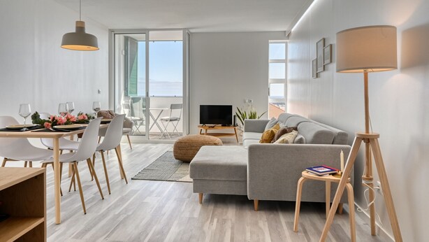 A modern living room with a grey sofa, dining table with chairs, a TV, and a lamp.