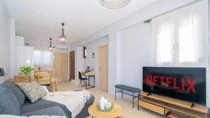 Un salon moderne avec un téléviseur à écran plat, un canapé gris, une table à manger en bois et un coin cuisine à l’arrière-plan.