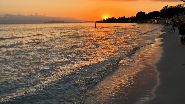 Ein Strand bei Sonnenuntergang, ruhiges Wasser und einige Personen, die am Ufer entlanggehen.