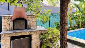 Ein Außenofen aus Stein zum Pizza-Braten mit Schornstein, umgeben von üppiger Vegetation und einem Swimmingpool.
