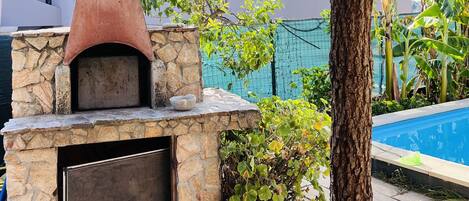 Un horno de piedra para pizzas al aire libre con chimenea, rodeado de abundante vegetación y una piscina.