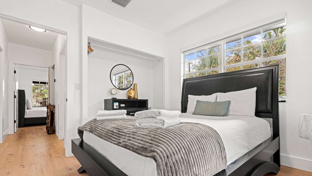 Een moderne slaapkamer met een groot bed, nachtkastje, spiegel en raam met uitzicht op bomen.