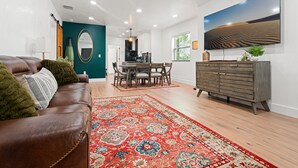 Een moderne woonkamer met een bruine leren bank, een vloerkleed met een rood patroon, een eethoek met een houten tafel en stoelen, en een grijs kastje met een flatscreen televisie aan de muur.