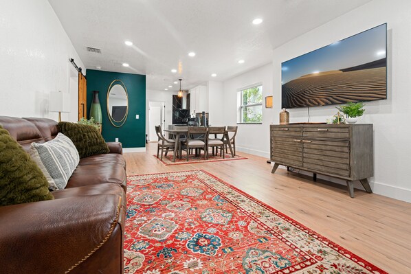 A modern living room with a brown leather sofa, a red patterned rug, a dining area with a wooden table and chairs, and a grey cabinet with a flat-screen TV mounted on the wall.