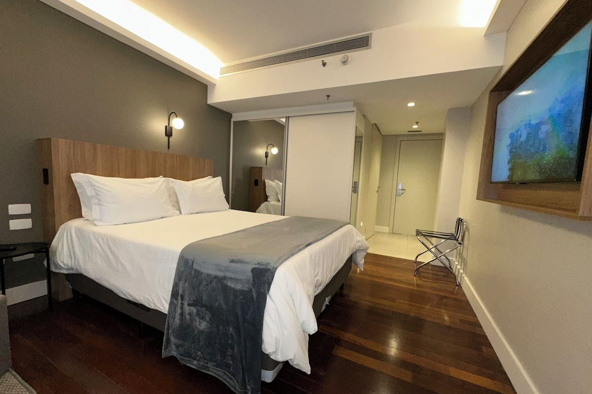 Una habitación de hotel moderna con una cama grande, una mesita de noche, un espejo, un televisor montado en la pared y una puerta que da al baño.