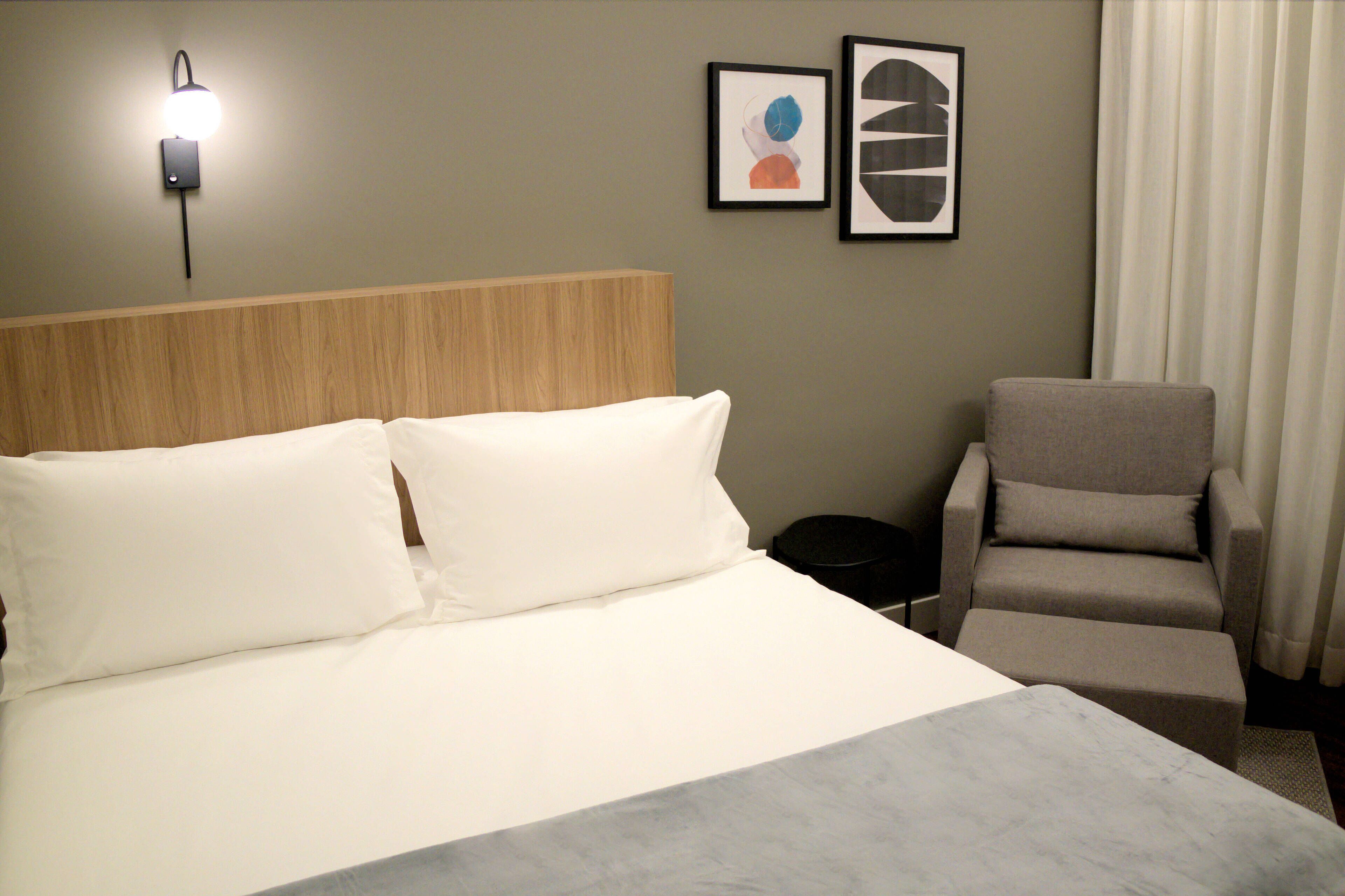 A hotel room with a bed, a chair, a wall-mounted light, and two framed artworks.