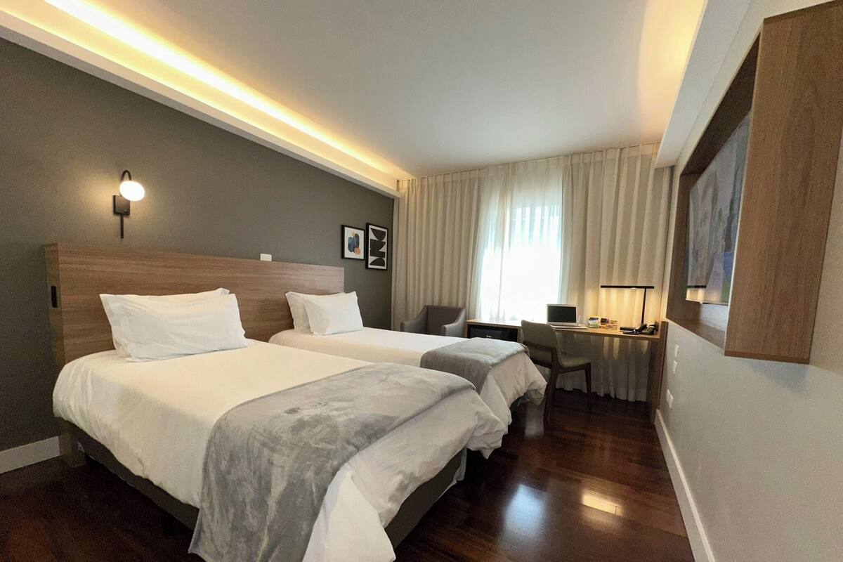 Habitación de hotel con dos camas, un escritorio con computadora, una silla y una ventana con cortinas.