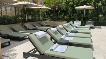 Filea di poltrone verdi con asciugamani e documenti, riparate da ombrelloni, in un cortile con uno sfondo verde e rigoglioso.