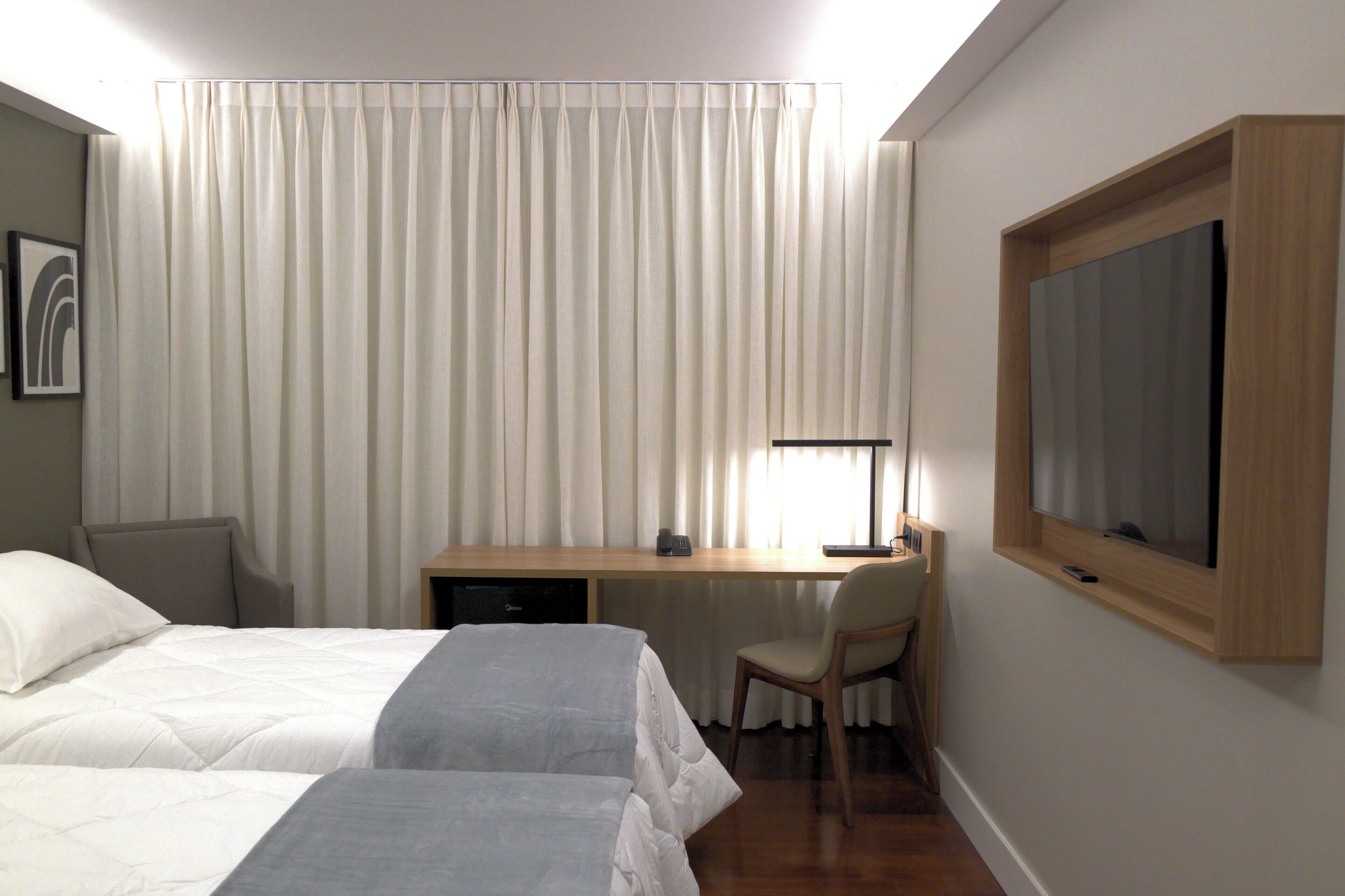 A hotel room with two beds, a desk, a chair, a television, and a window with curtains.