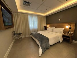 Una camera d'albergo con un letto grande, una TV fissata al muro, una poltrona e un comodino con una lampada.