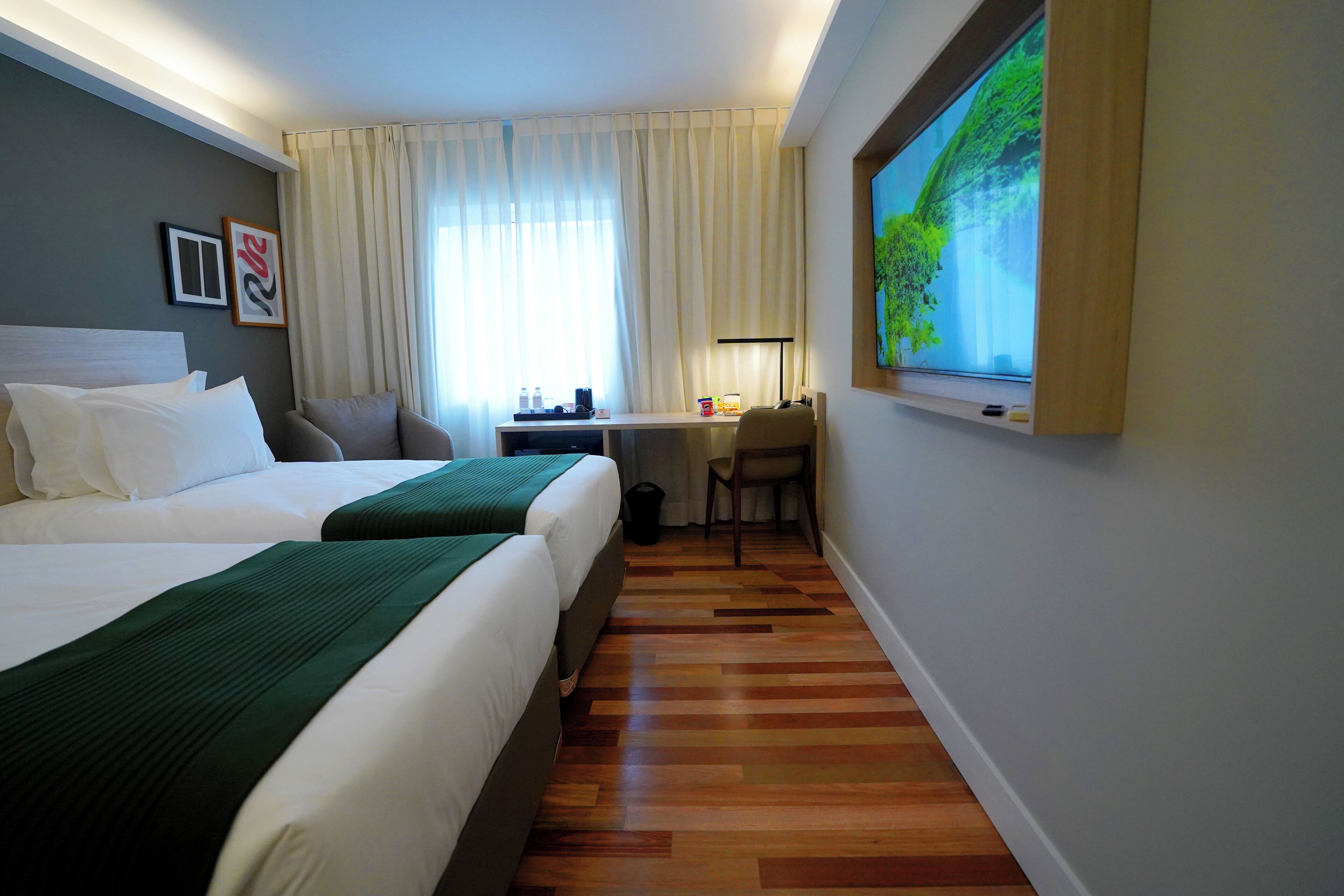 Ein Hotelzimmer mit zwei Betten, einem Schreibtisch, einem Stuhl, einem Fernseher und einem großen Fenster mit Vorhängen.