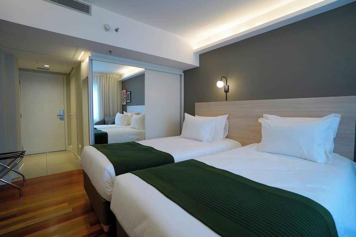 Habitación de hotel con dos camas, un espejo, una mesita de noche y una lámpara de pared.