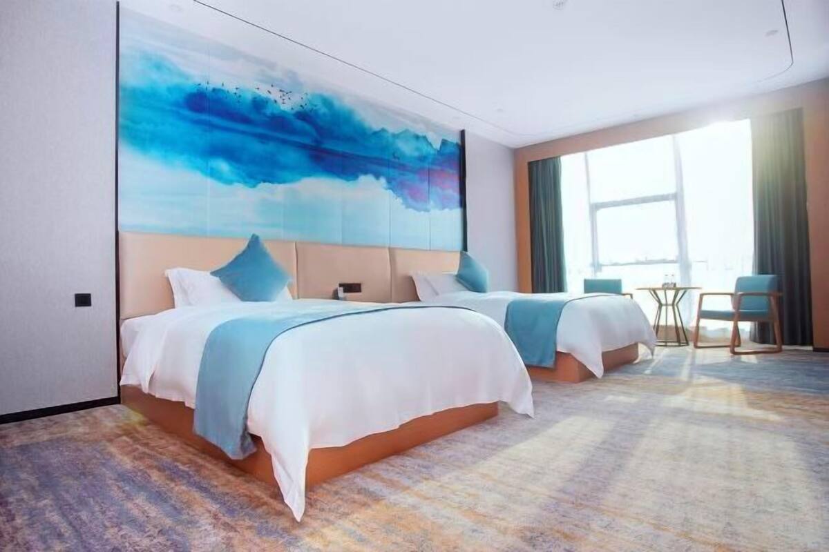 A hotel room with two beds, a large abstract painting, a view of the city, and a small table with chairs.