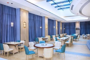 Espaço de refeições de hotel com mesas redondas, cadeiras e cortinas azuis.