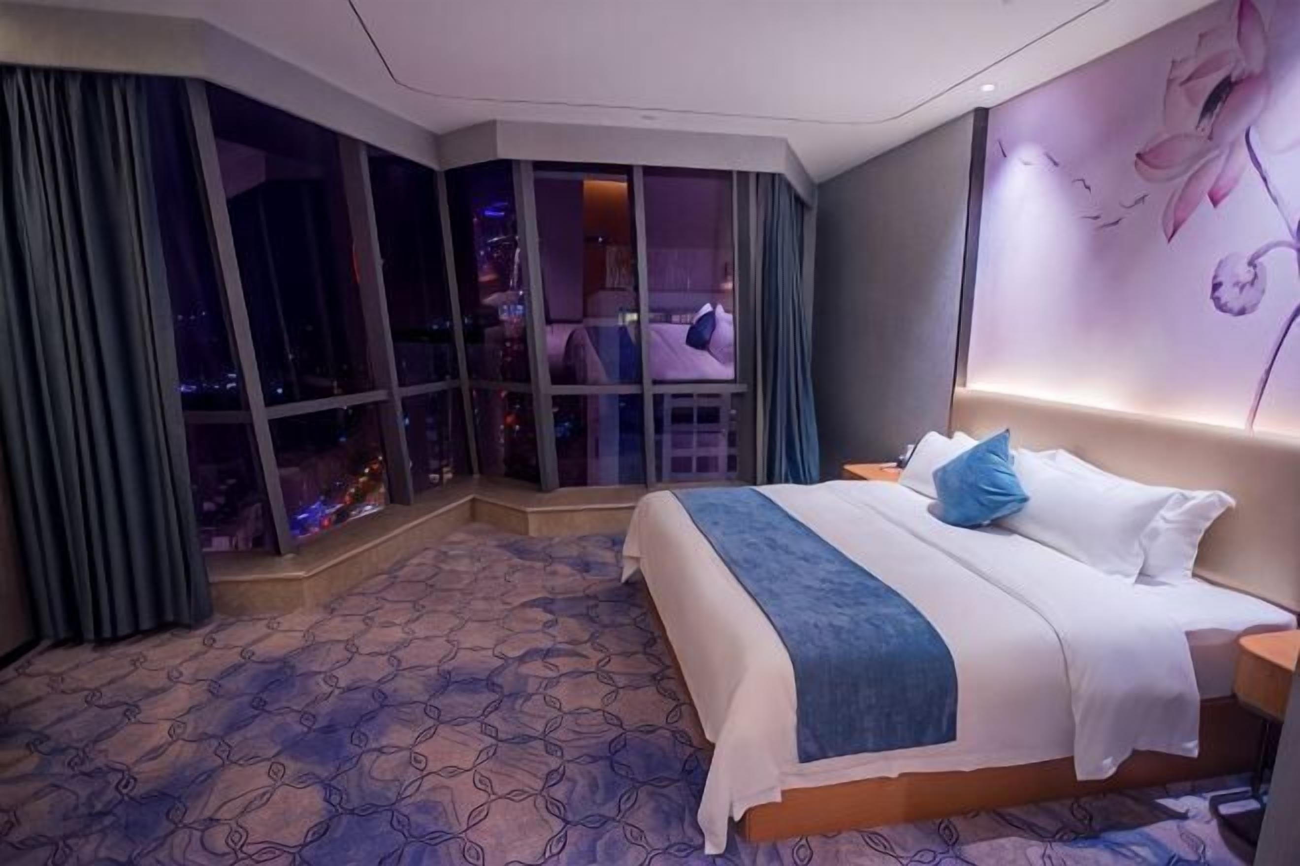 A hotel room with a large bed, a city view through floor-to-ceiling windows, a patterned carpet, and a wall with a floral design.