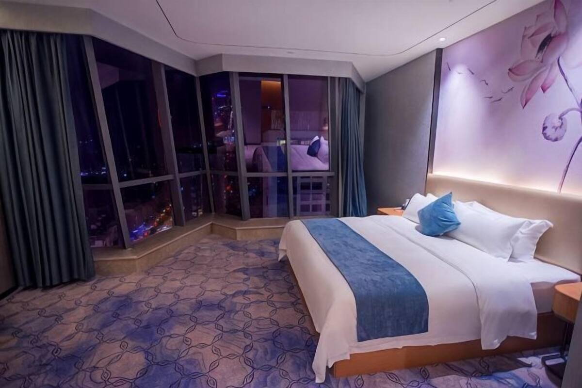 A hotel room with a large bed, a city view through floor-to-ceiling windows, a patterned carpet, and a wall with a floral design.
