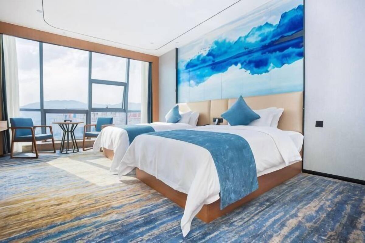 A hotel room with two beds, a large window, a view of the mountains, and a blue abstract painting on the wall.