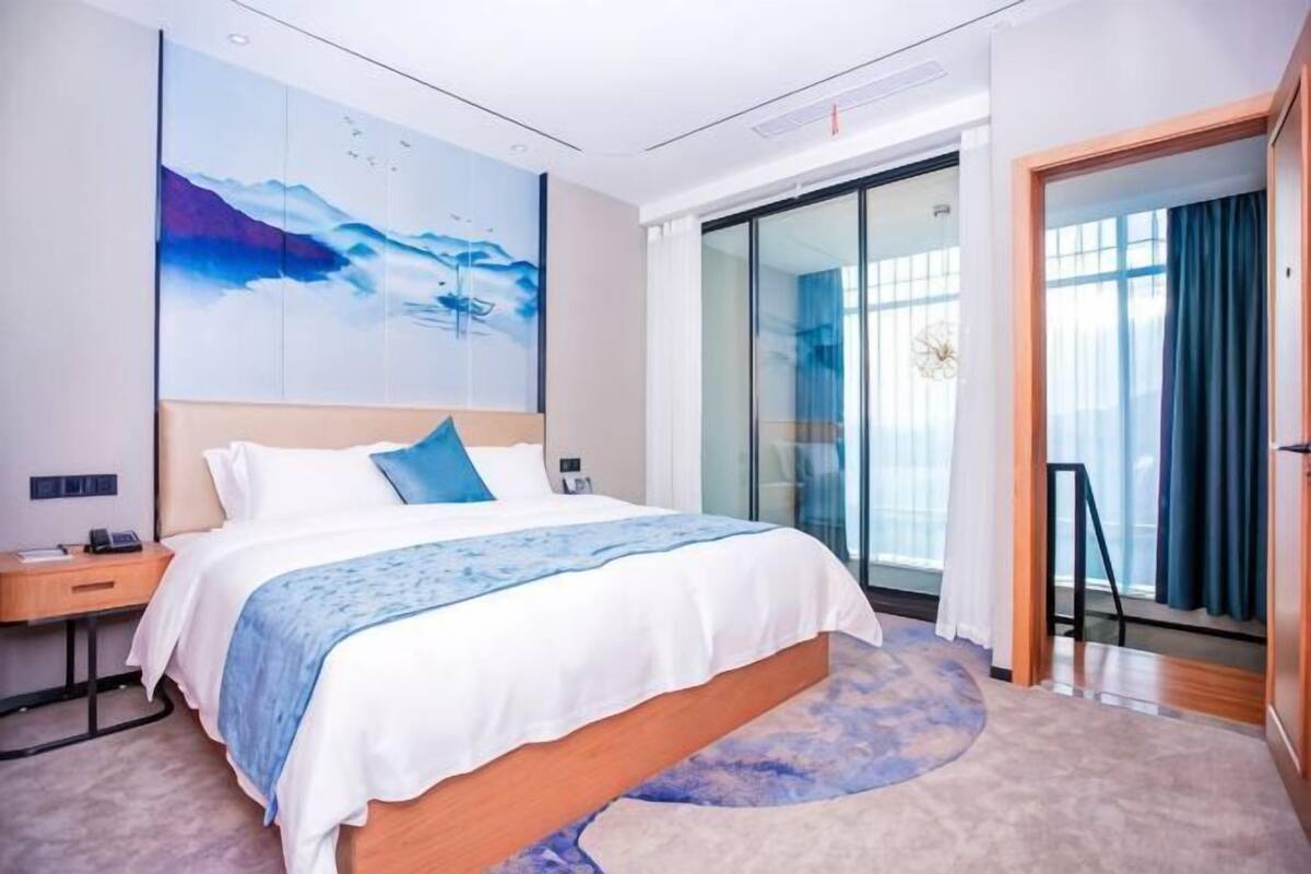 A modern hotel room with a large bed, a nightstand, a wall-mounted artwork, and a sliding glass door leading to a balcony.
