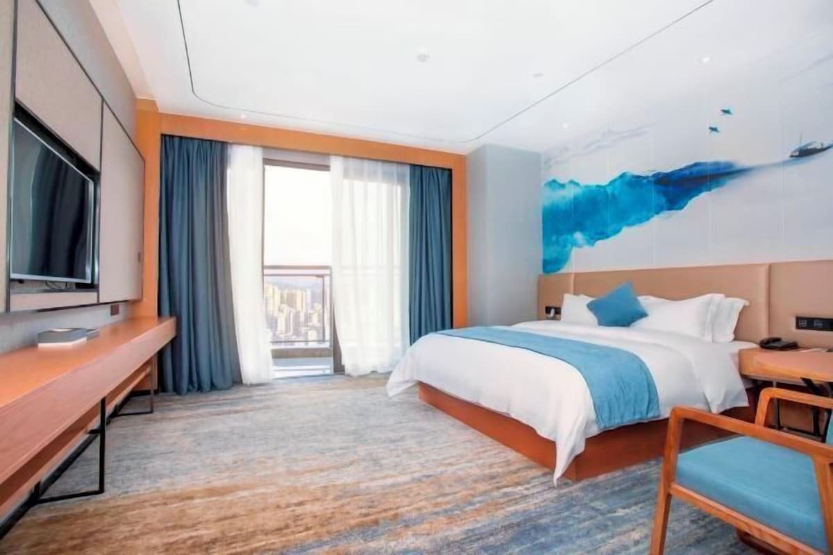 A modern hotel room with a large bed, a desk, a television, and a balcony with curtains.