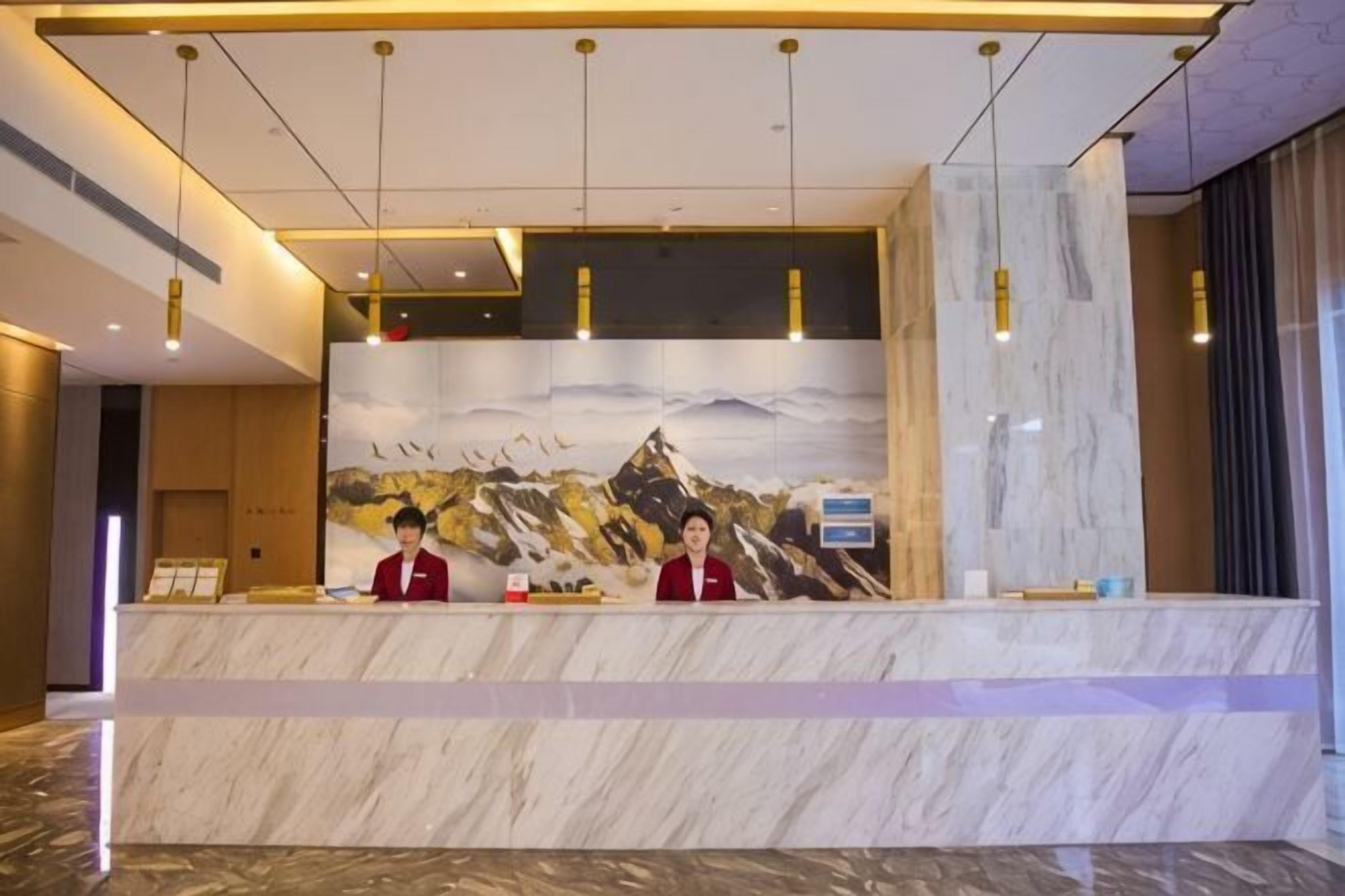 Un mostrador de recepción con encimera de mármol, dos recepcionistas con uniformes rojos y un gran mural de montañas en la pared.