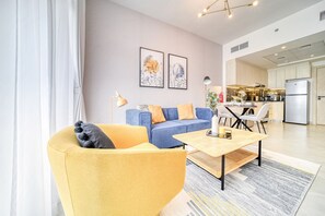 A modern living room with a sofa, armchair, coffee table, and a dining area with chairs. There is a kitchen visible in the background.