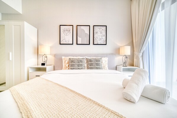 A hotel room with a large bed, two bedside tables with lamps, a wall with three framed artworks, and a window with curtains.