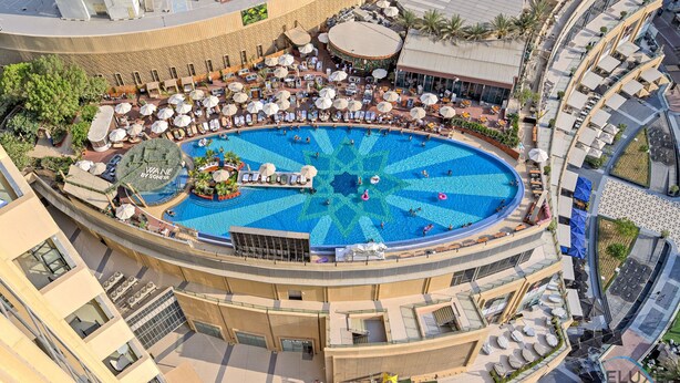Une grande piscine circulaire, entourée de plusieurs chaises longues et de parasols, et bordée d’un bâtiment aux lignes courbes, doté de balcons et d’espaces de détente en plein air.