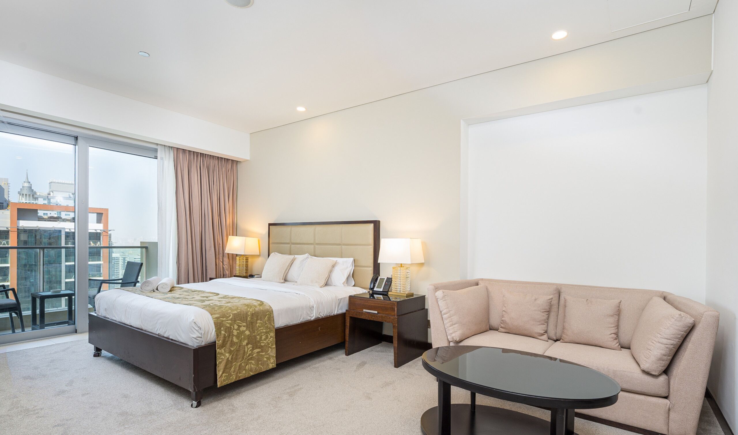 A hotel room with a bed, bedside tables, a sofa, a glass table, and a view of the city.