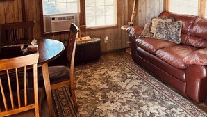 A rustic interior with wooden walls, a ceiling fan, a leather couch, a dining table with chairs, and large windows with blinds. - Prairie Creek Cabin -Ride SxS to trails from the cabin! Kayak and Fish the Creek (Mena)