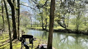 A camping chair with fishing gear by a calm river in a wooded area. - Prairie Creek Cabin -Ride SxS to trails from the cabin! Kayak and Fish the Creek (Mena)