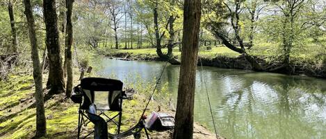 A camping chair with fishing gear by a calm river in a wooded area.