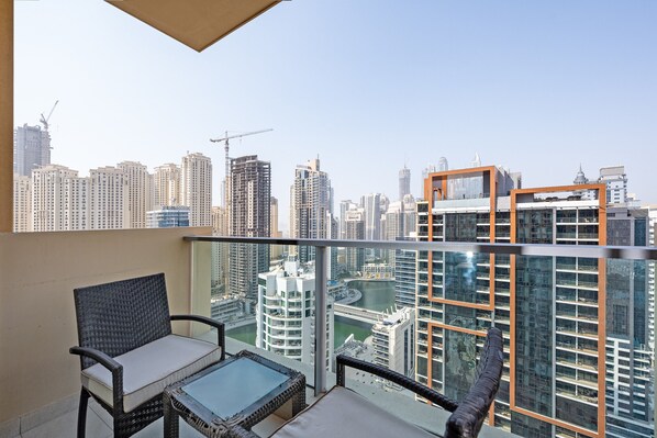 A balcony with wicker furniture and a glass table overlooking a cityscape with high-rise buildings and a river.