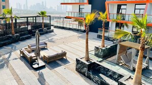 A rooftop terrace with outdoor seating, palm trees, and a city skyline in the background. - Homely 1BR at Binghatti Avenue Al Jaddaf by Deluxe Holiday Homes (Dubai)