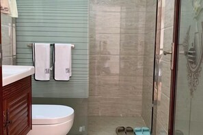 A bathroom with a toilet, two towels hanging on a wall, a shower area, and a pair of slippers on the floor.
