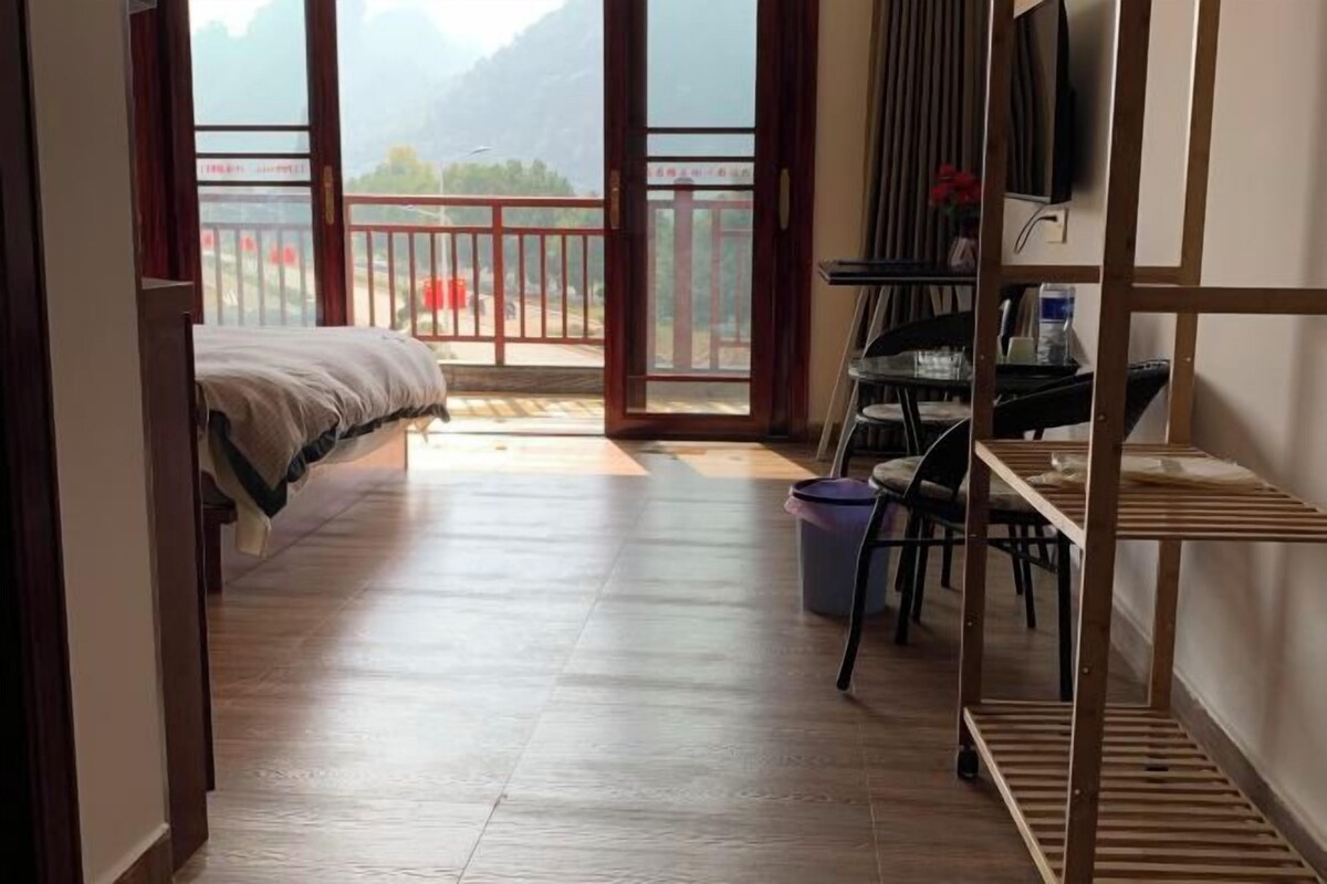 A room with a bed, a desk, a chair, a TV, and a view of mountains through a balcony door.