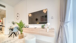 Uma sala de estar moderna com uma grande televisão de tela plana na parede, uma área de jantar com mesa e cadeiras, e uma cozinha visível ao fundo.