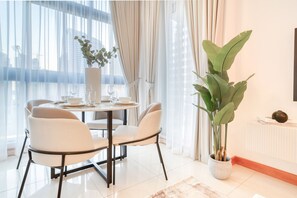 Un espace repas moderne avec une table ronde, des chaises blanches, une plante en pot et une vue sur la ville grâce à de grandes fenêtres.