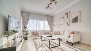 A modern living room with a sofa, coffee table, and a TV on a stand. There are plants, a rug, and curtains.