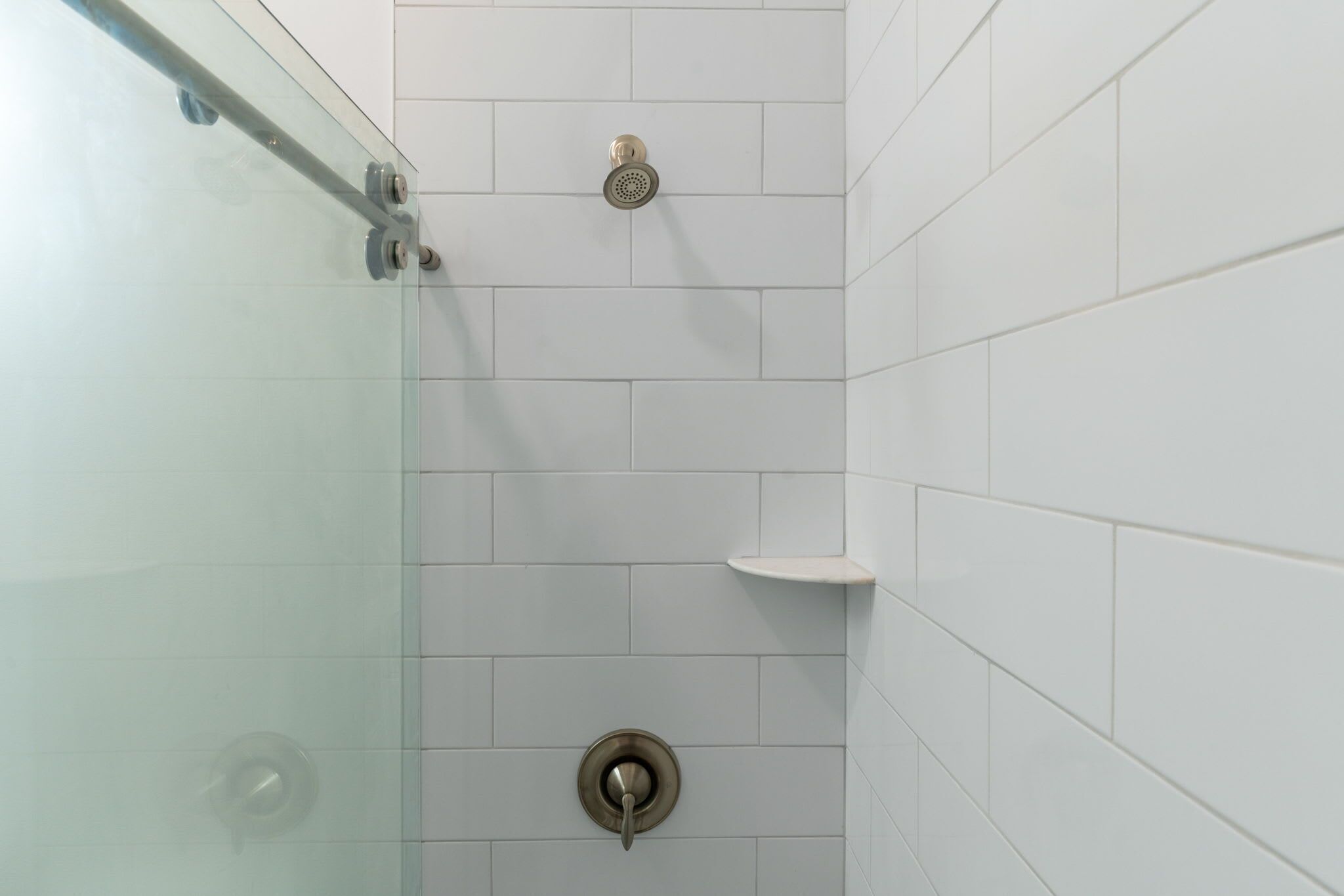 Ducha con puerta de cristal, paredes de azulejos blancos y un estante pequeño.