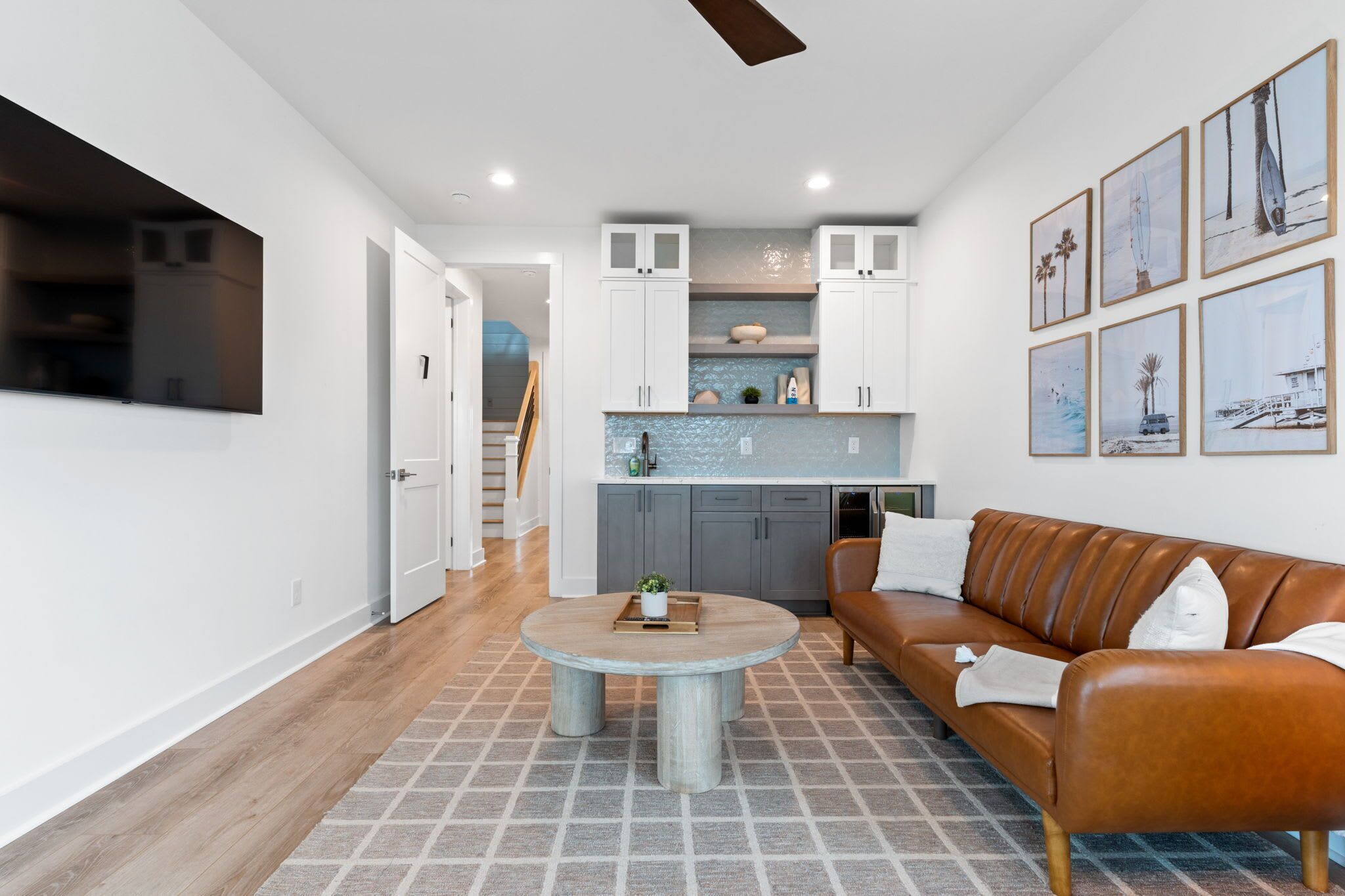 A modern living room with a brown leather sofa, a round coffee table, and a kitchen area with white cabinets and a blue backsplash.