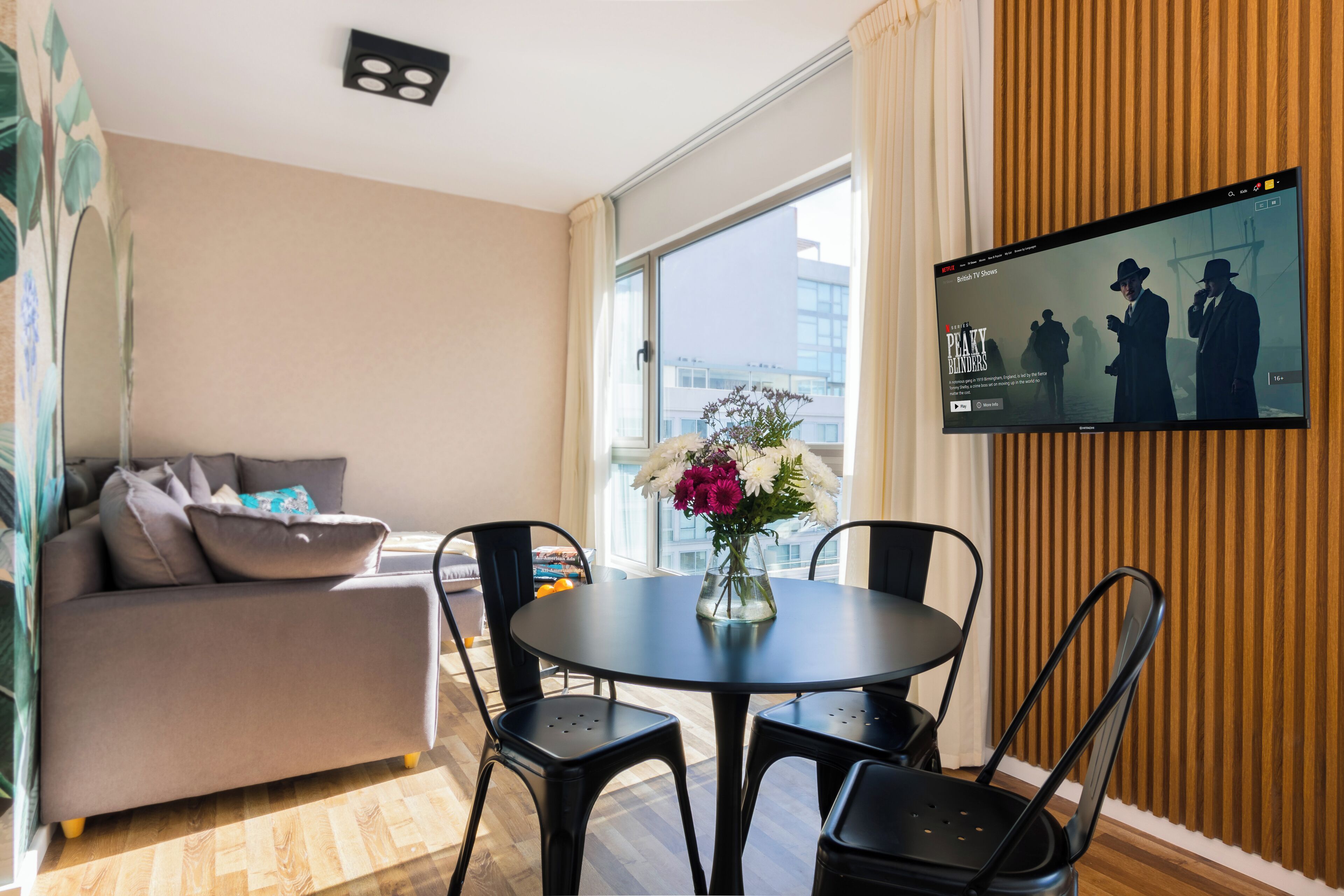 A modern living room with a sofa, a round table with chairs, a TV on the wall, and a window with a view of buildings.