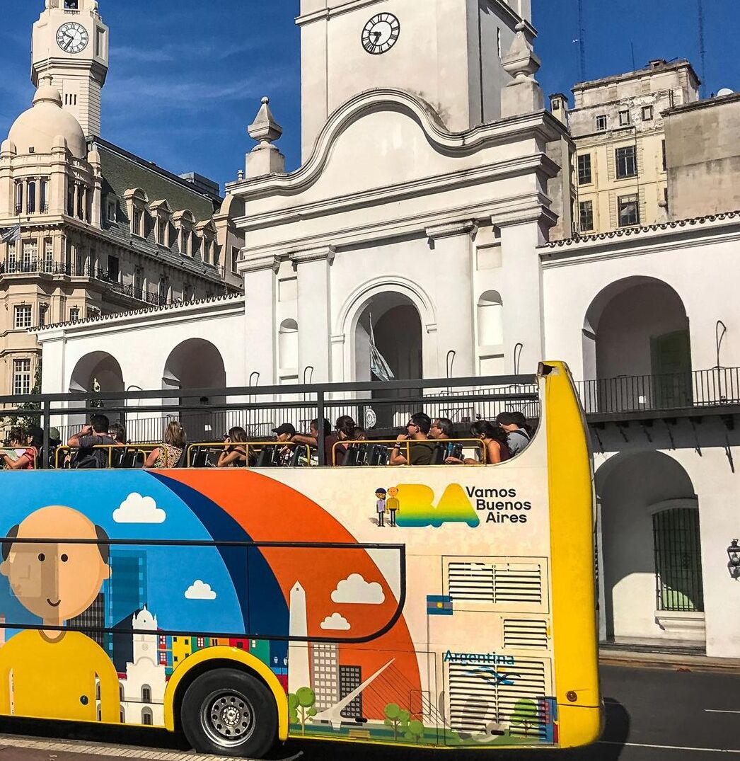 A colorful bus with "Vamos Buenos Aires" on the side, parked in front of a historic building with arches and a clock tower.