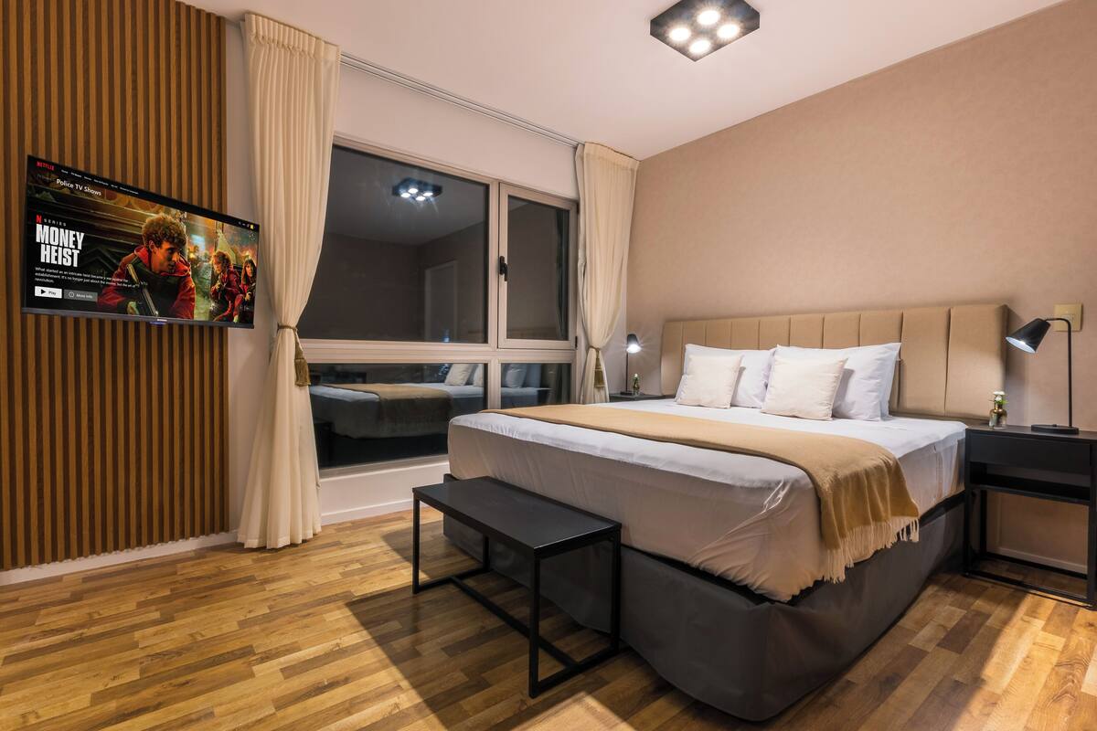 Une chambre à coucher moderne avec un grand lit, un téléviseur fixé au mur, une table de chevet et une fenêtre avec des rideaux.